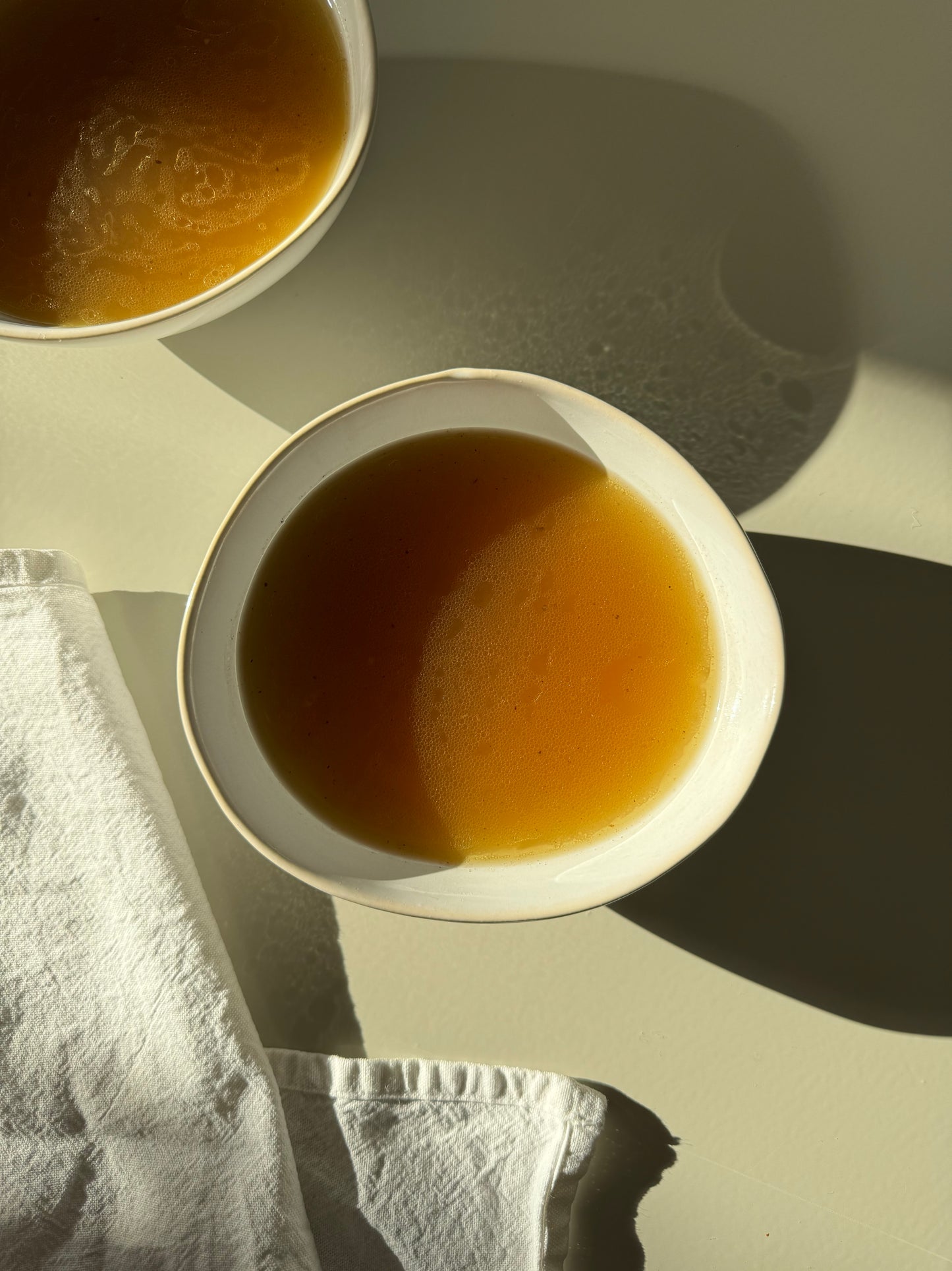 Two bowls of broth on a light surface, with one bowl partially covered by a white napkin.