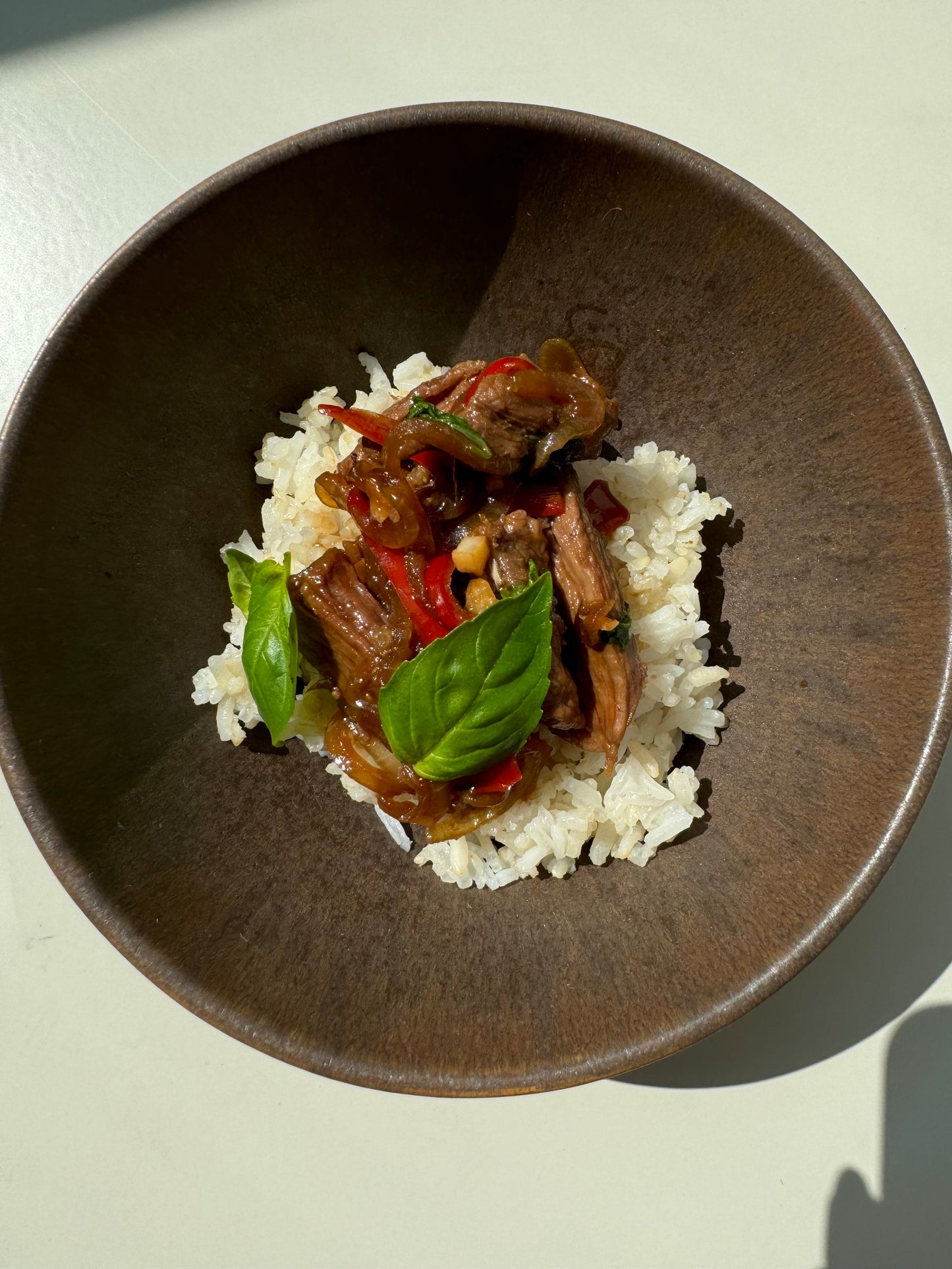 A bowl of Basil & Beef Strips Congee, with the congee having chunks of beef, basil leaves, and a dash of coconut sugar, served on a white plate against a light background.