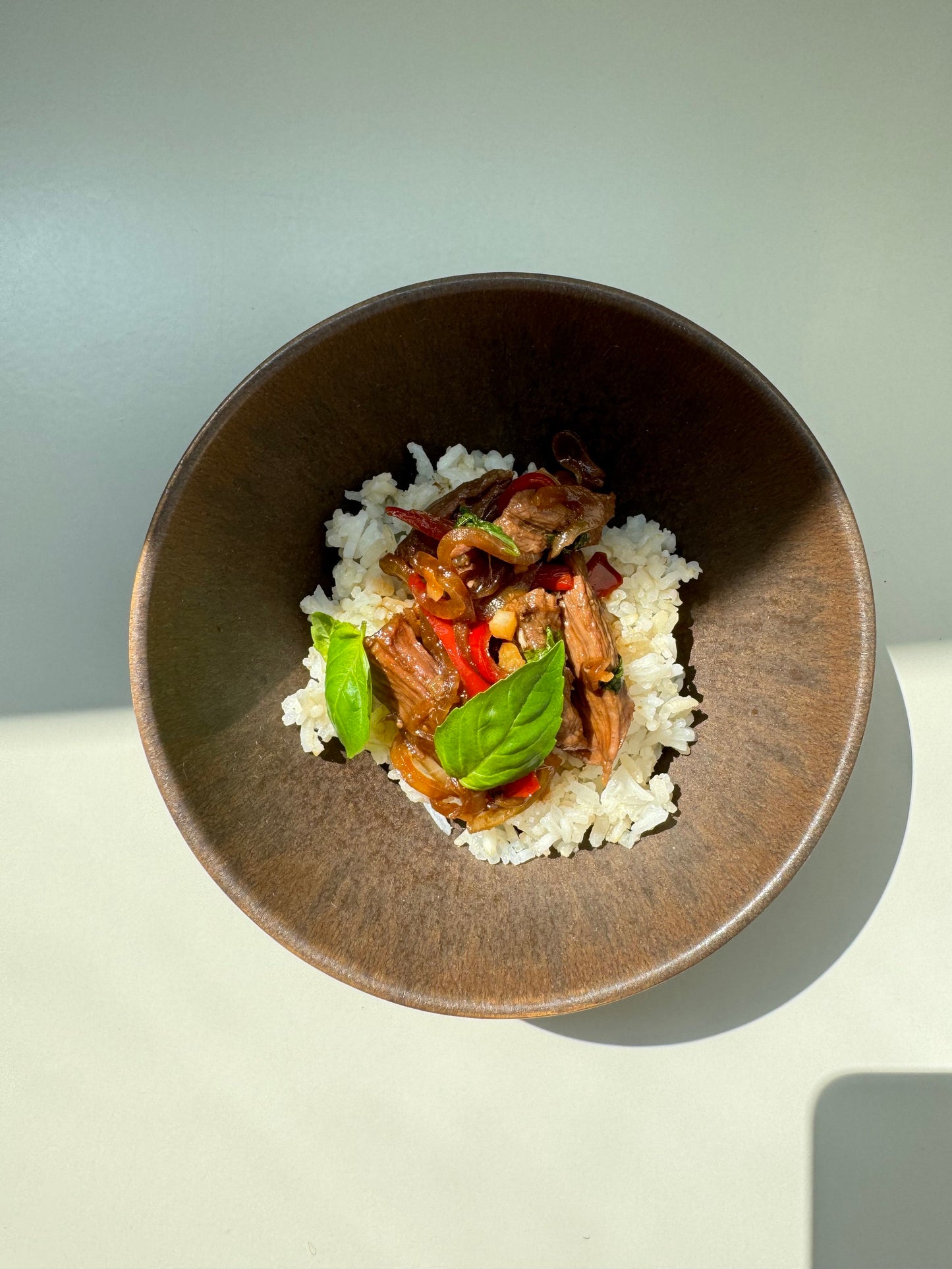 A bowl of Basil & Beef Strips Congee, with the congee having chunks of beef, basil leaves, and a dash of coconut sugar, served on a white plate against a light background.