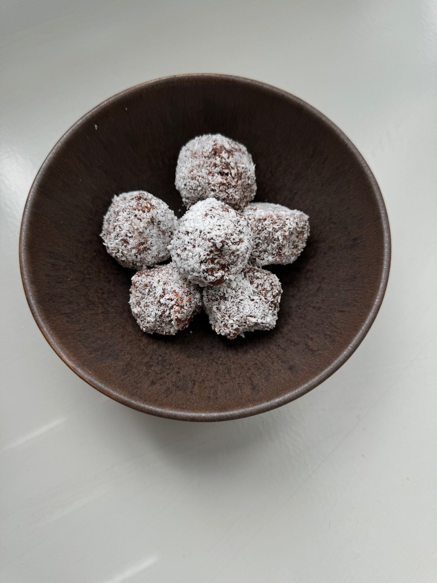 A bowl filled with cacao and dates bliss balls, with a textured brown rim and a shiny white surface.