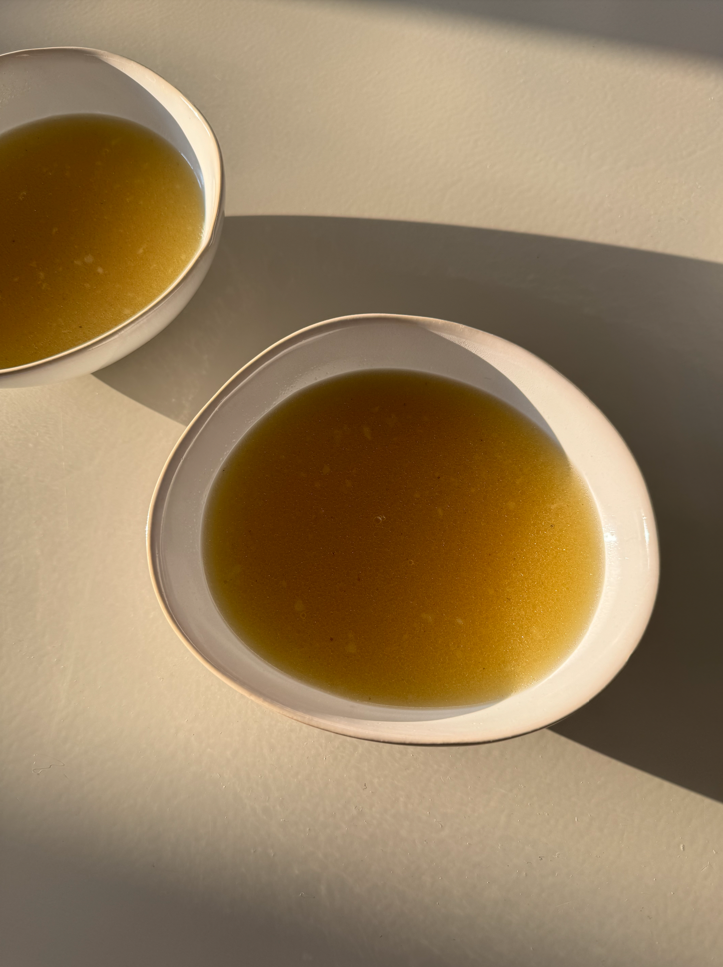 Two white bowls filled with chicken broth, placed on a light-colored surface, possibly representing a breakfast or brunch setting.