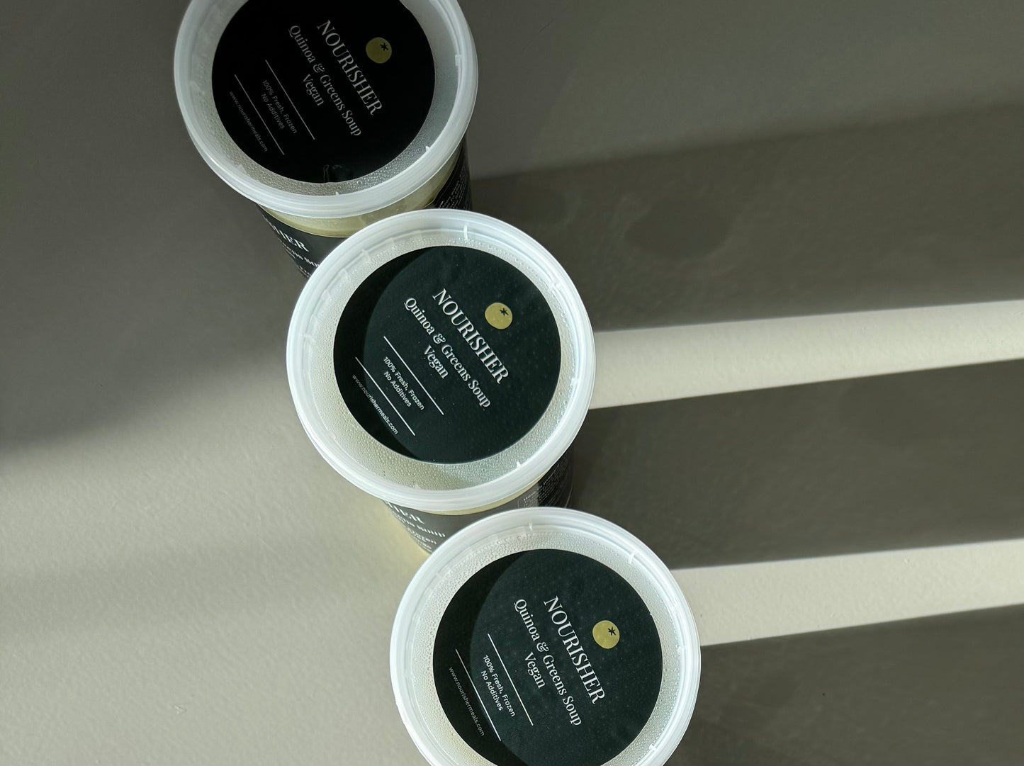 packaged units of creamy Quinoa Greens soup next to a scattering of red lentils on a light surface, with a shadow falling over part of the image.