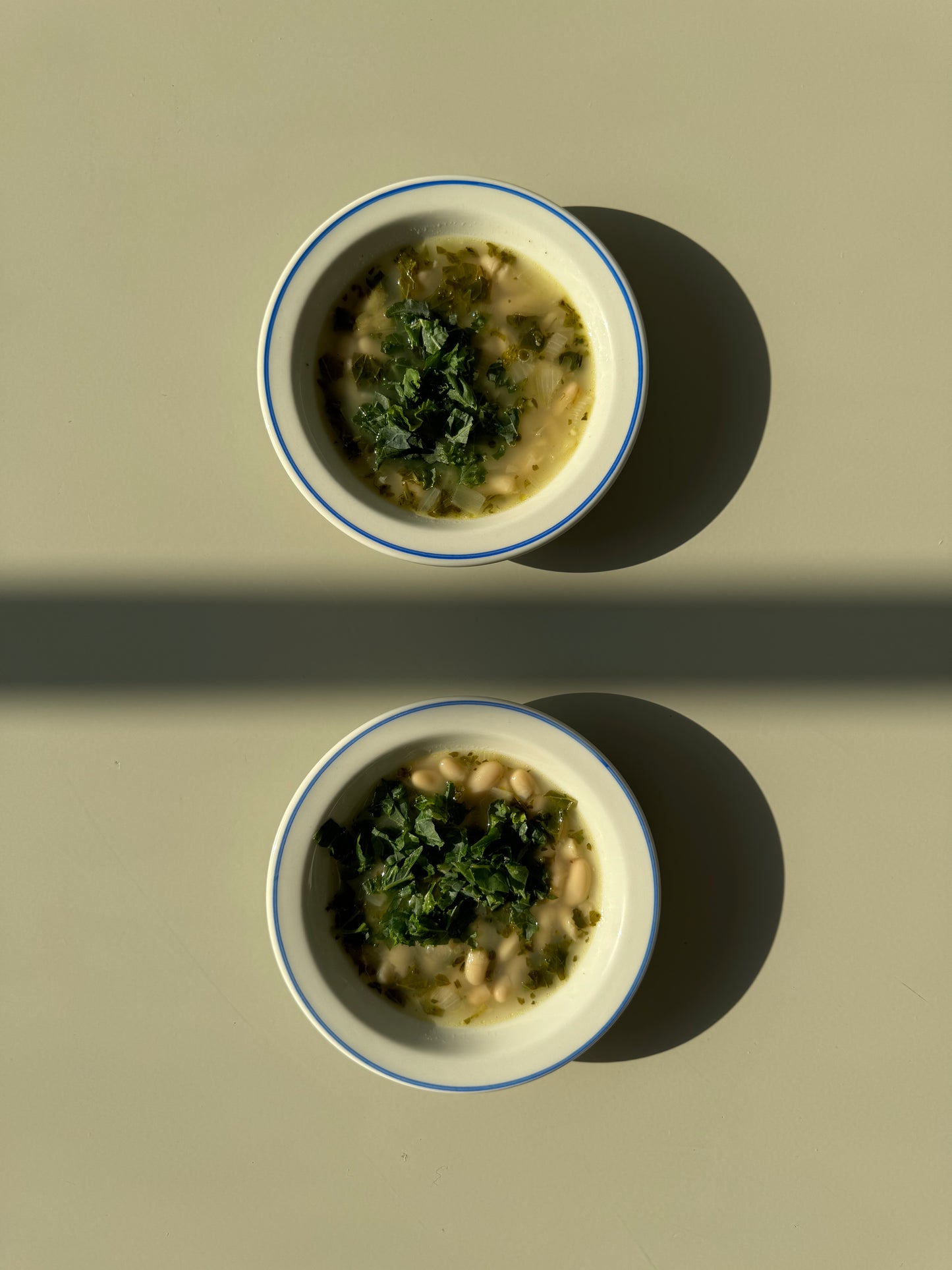 A bowl of Lemony Kale and White Bean Soup on a plate with lemons beside it, presented on a white background with shadows.