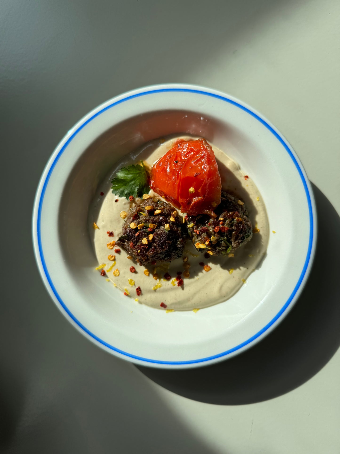 A dish containing kofta (meatballs) on tahini sauce, garnished with fresh parsley, served with a side of tomato.