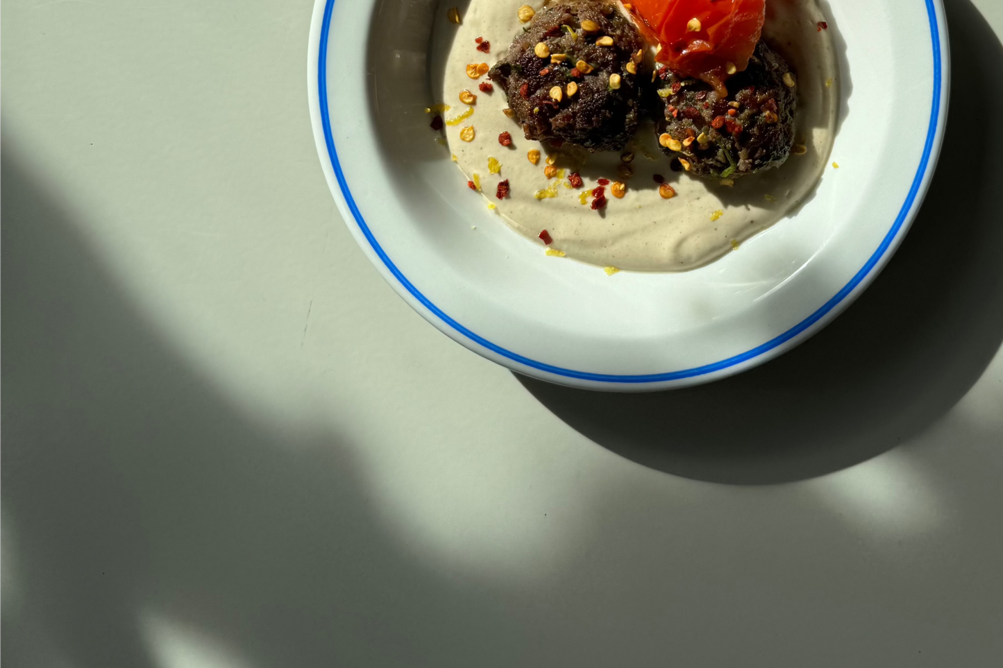 A dish containing kofta (meatballs) on tahini sauce, garnished with fresh parsley, served with a side of tomato.