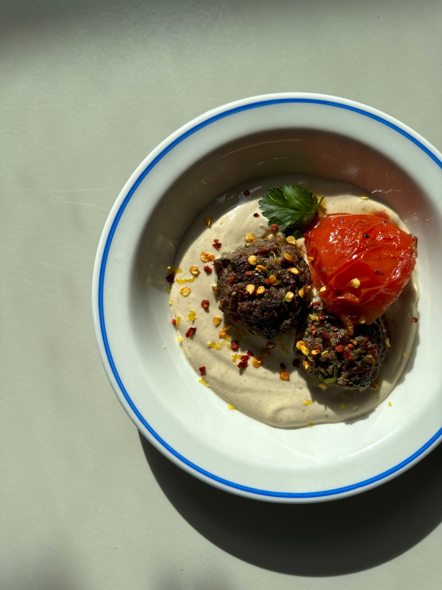 A dish containing kofta (meatballs) on tahini sauce, garnished with fresh parsley, served with a side of tomato.