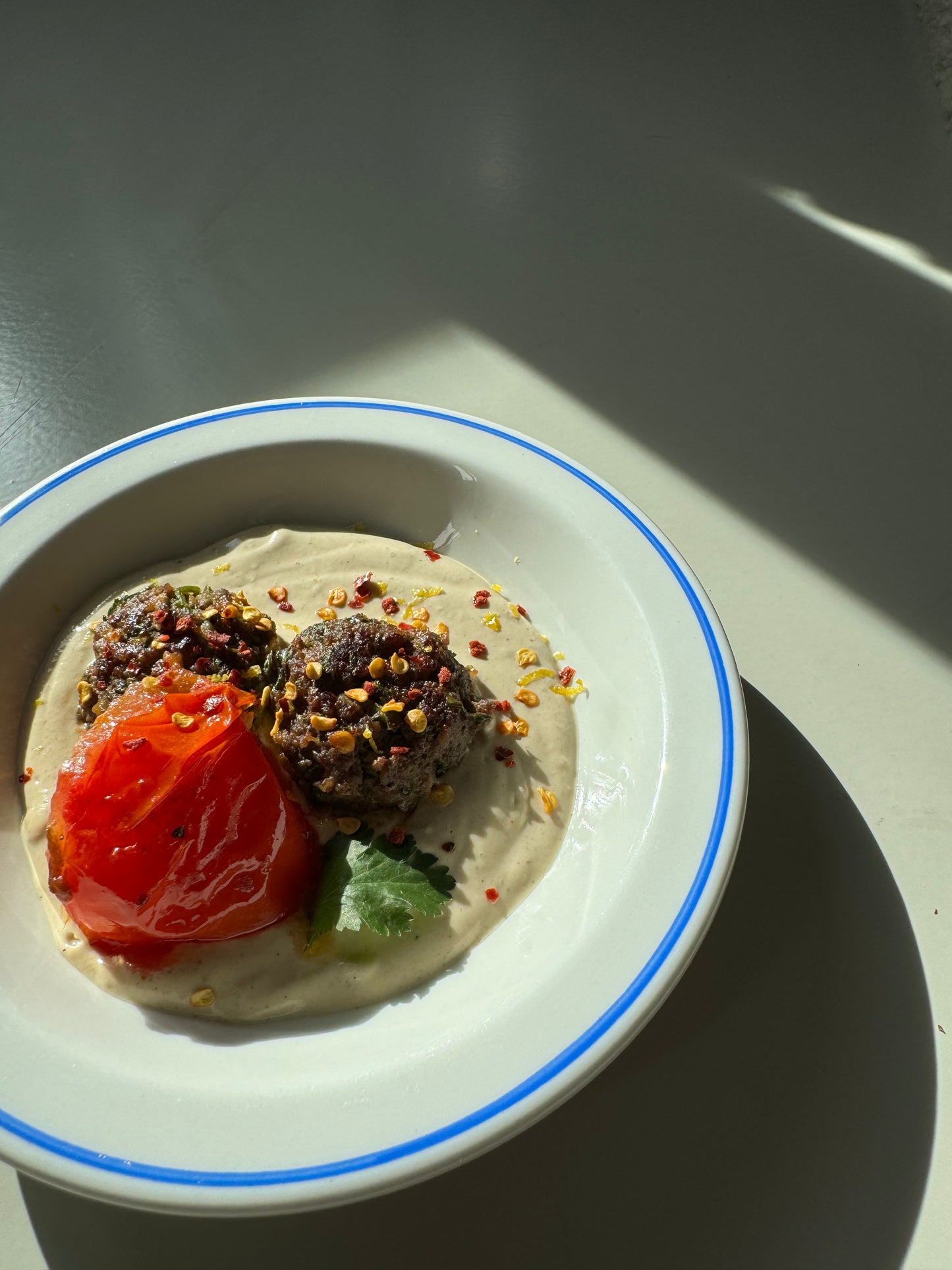 A dish containing kofta (meatballs) on tahini sauce, garnished with fresh parsley, served with a side of tomato.