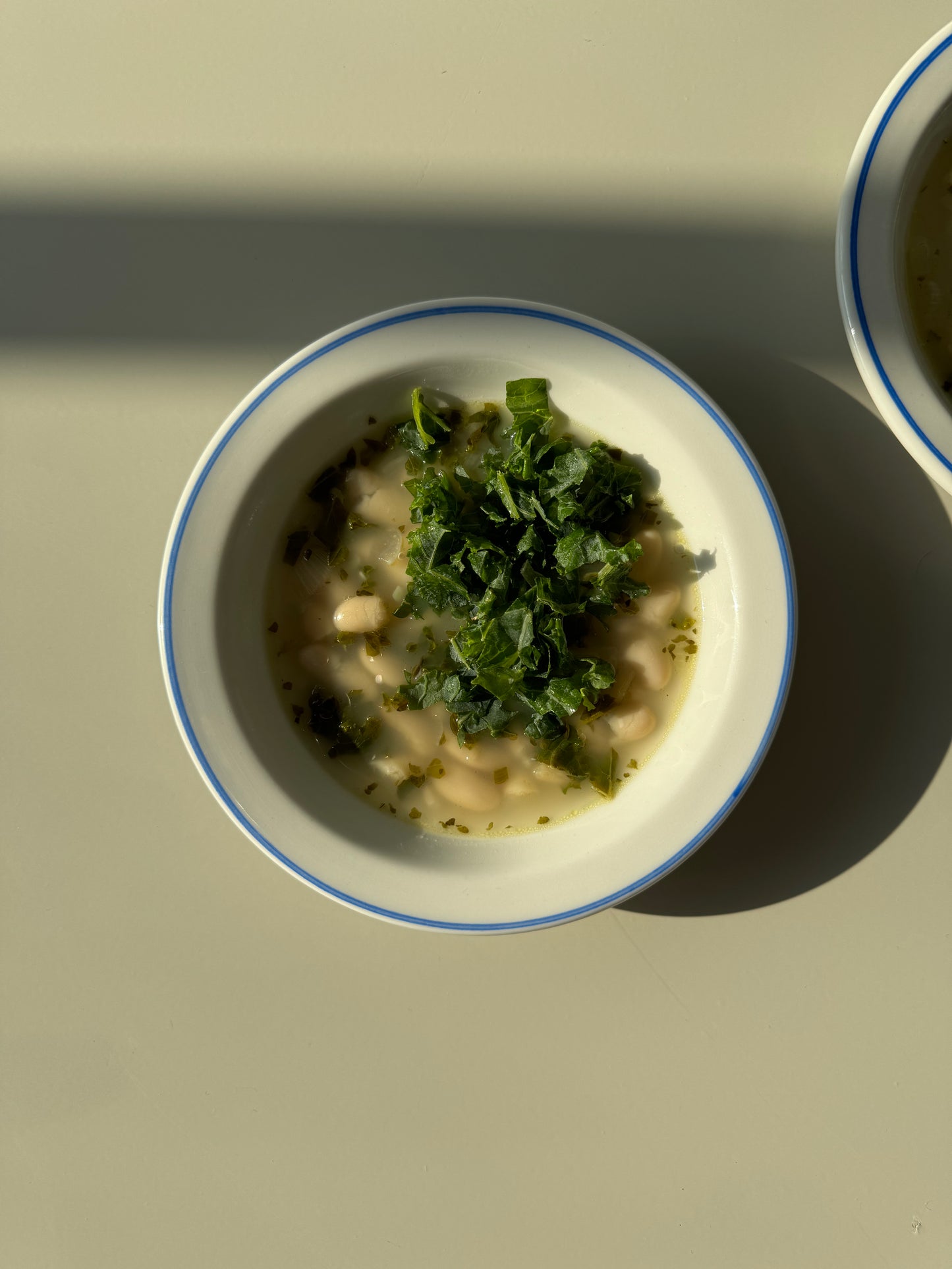 A bowl of Lemony Kale and White Bean Soup on a plate with lemons beside it, presented on a white background with shadows.