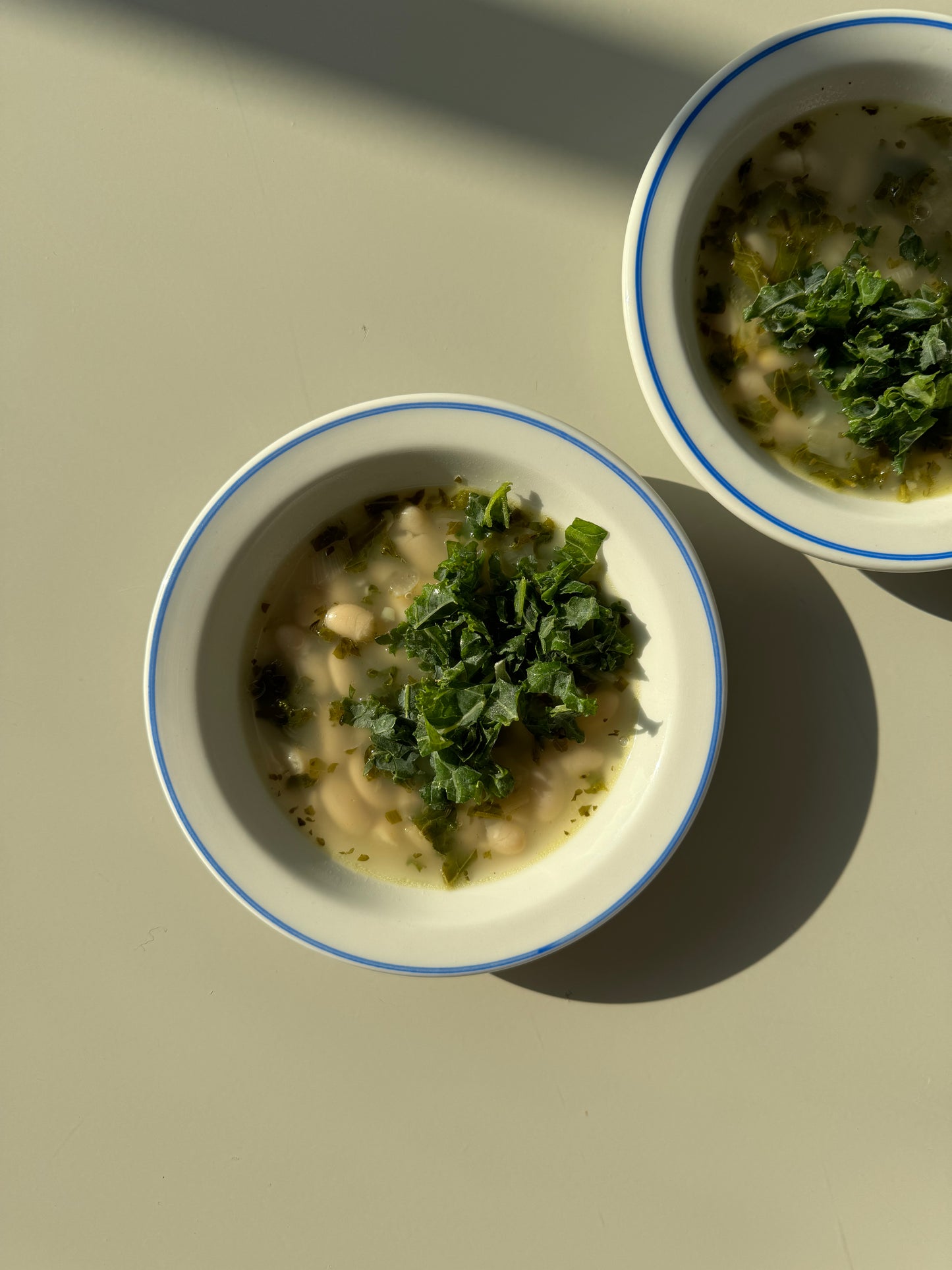 A bowl of Lemony Kale and White Bean Soup on a plate with lemons beside it, presented on a white background with shadows.