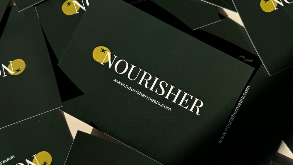 A pile of dark green gift cards with the word 'NOURISHER' printed in white, arranged in a casual pattern.