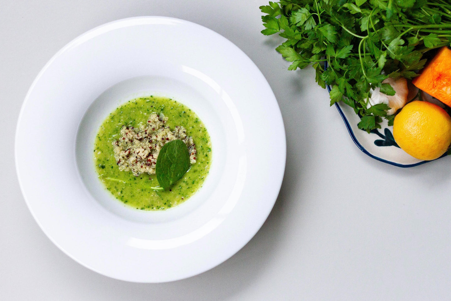 A bowl of creamy Quinoa Greens soup next to a scattering of red lentils on a light surface, with a shadow falling over part of the image.