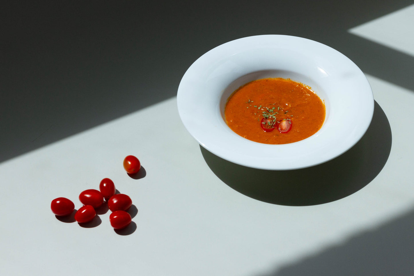 A bowl of roasted tomato soup with a garnish of basil leaves on a white plate, with a shadow falling over the plate.