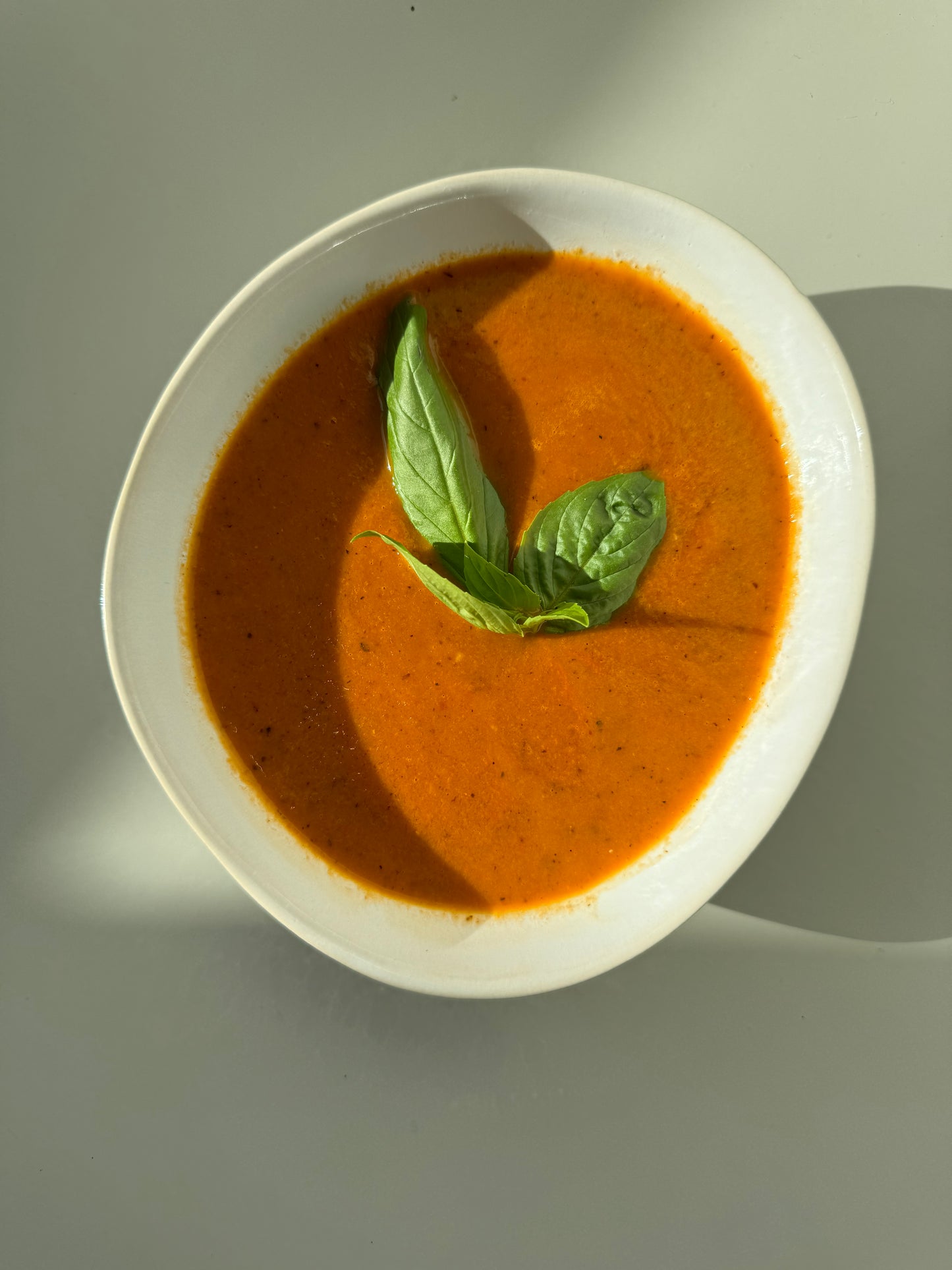 A bowl of roasted tomato soup with a garnish of basil leaves on a white plate, with a shadow falling over the plate.
