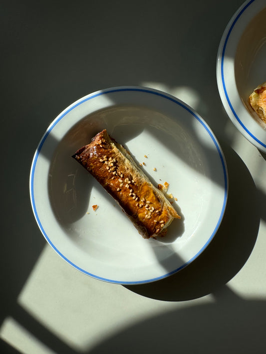 A single borek with feta cheese and parsley on a round white plate with a blue rim, placed on a surface with a shadow pattern.