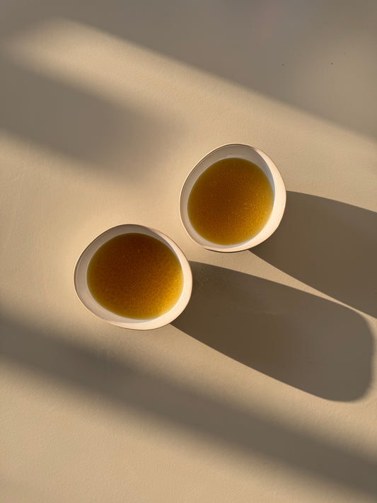 Two white bowls filled with chicken broth, placed on a light-colored surface, possibly representing a breakfast or brunch setting.