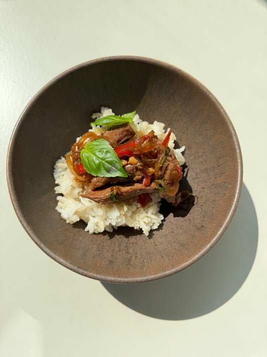 A bowl of Basil & Beef Strips Congee, with the congee having chunks of beef, basil leaves, and a dash of coconut sugar, served on a white plate against a light background.
