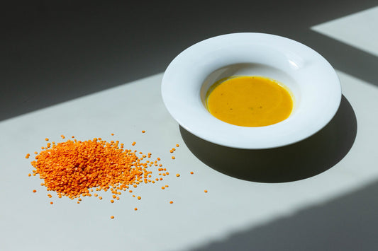 A bowl of creamy squash and red lentils soup next to a scattering of red lentils on a light surface, with a shadow falling over part of the image.