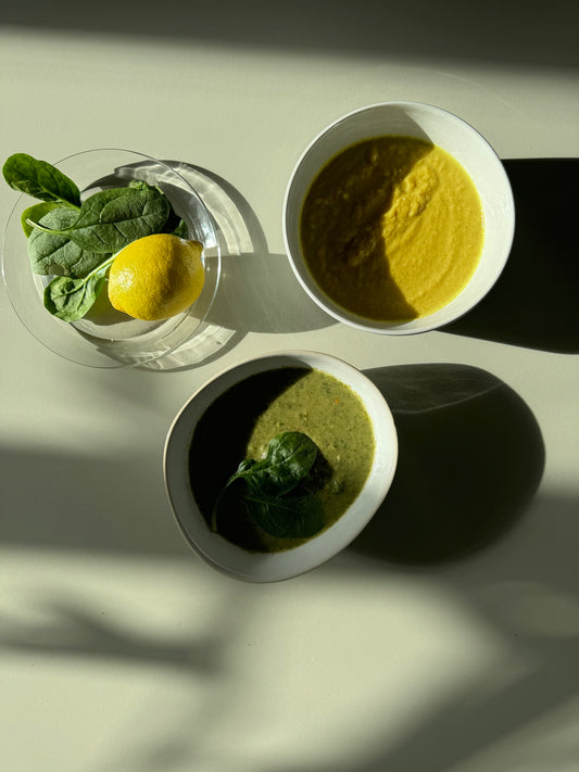 Two bowls of soup, one with broccoli and peas, the other with just spinach and a lemon slice, on a surface with a shadow falling over part of the image.