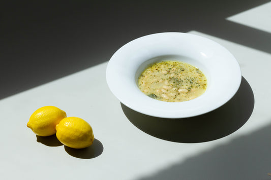 A bowl of Lemony Kale and White Bean Soup on a plate with lemons beside it, presented on a white background with shadows.