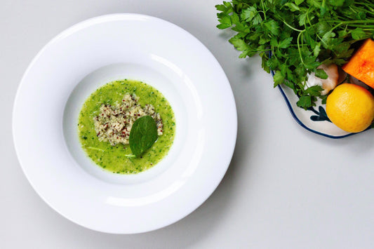 A bowl of creamy Quinoa Greens soup next to a scattering of red lentils on a light surface, with a shadow falling over part of the image.