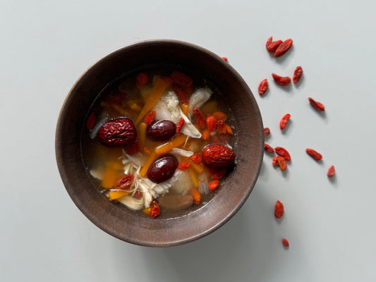 A bowl of Chicken, Red Dates & Ginger Soup with the soup still hot and red dates and ginger slices visible in the bowl, on a light background.