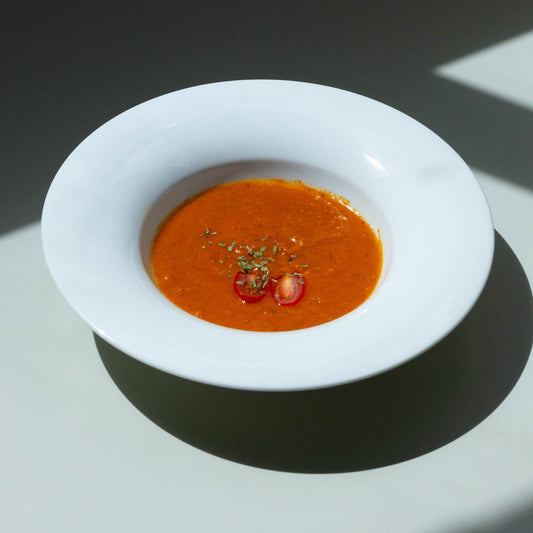 A bowl of roasted tomato soup with a garnish of basil leaves on a white plate, with a shadow falling over the plate.